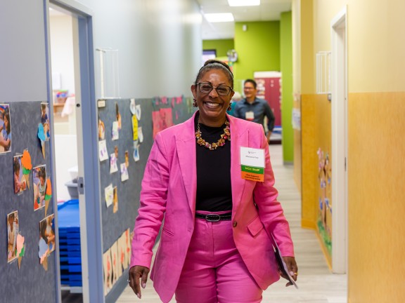 Marianne Fray, President and CEO, walks down a hallway at the childcare center, smiling widely and wearing a pink suit, glasses, and a nametag