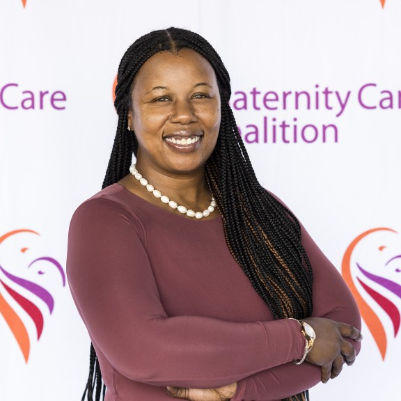 Aisha Brathwaite standing in front of MCC backdrop with folded arms, wearing a maroon longsleeve top and a white beaded necklace, and smiling. 