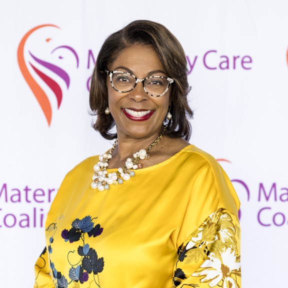 Marianne Fray stands smiling in front of MCC backdrop in yellow shirt, wearing glasses, and smiling.