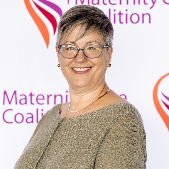 Karen Pollack standing in front of MCC backdrop, wearing a brown sweater, glasses, and smiling.