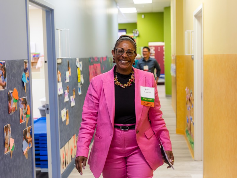 Marianne Fray, President and CEO, walks down a hallway at the childcare center, smiling widely and wearing a pink suit, glasses, and a nametag